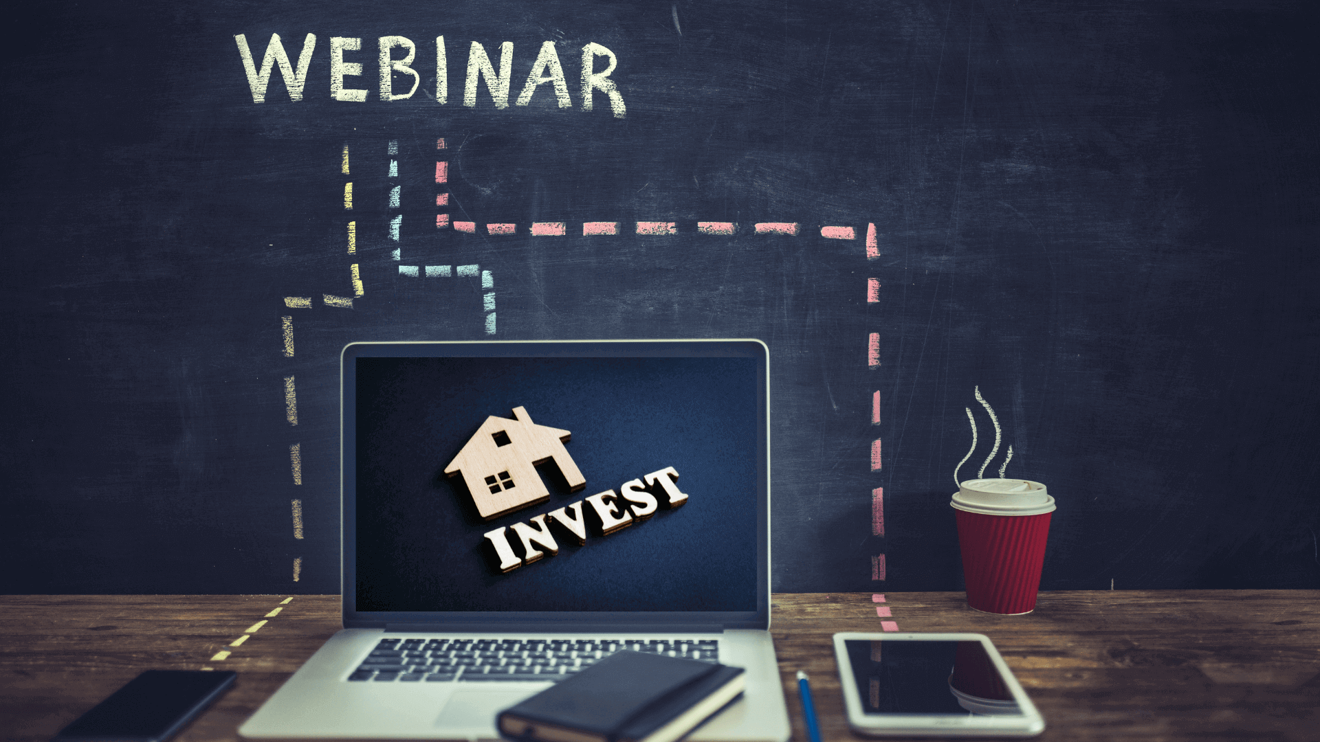 Laptop mit dem Wort 'INVEST' und einem Haus-Symbol auf dem Bildschirm, daneben ein Kaffeebecher und ein Notizbuch.